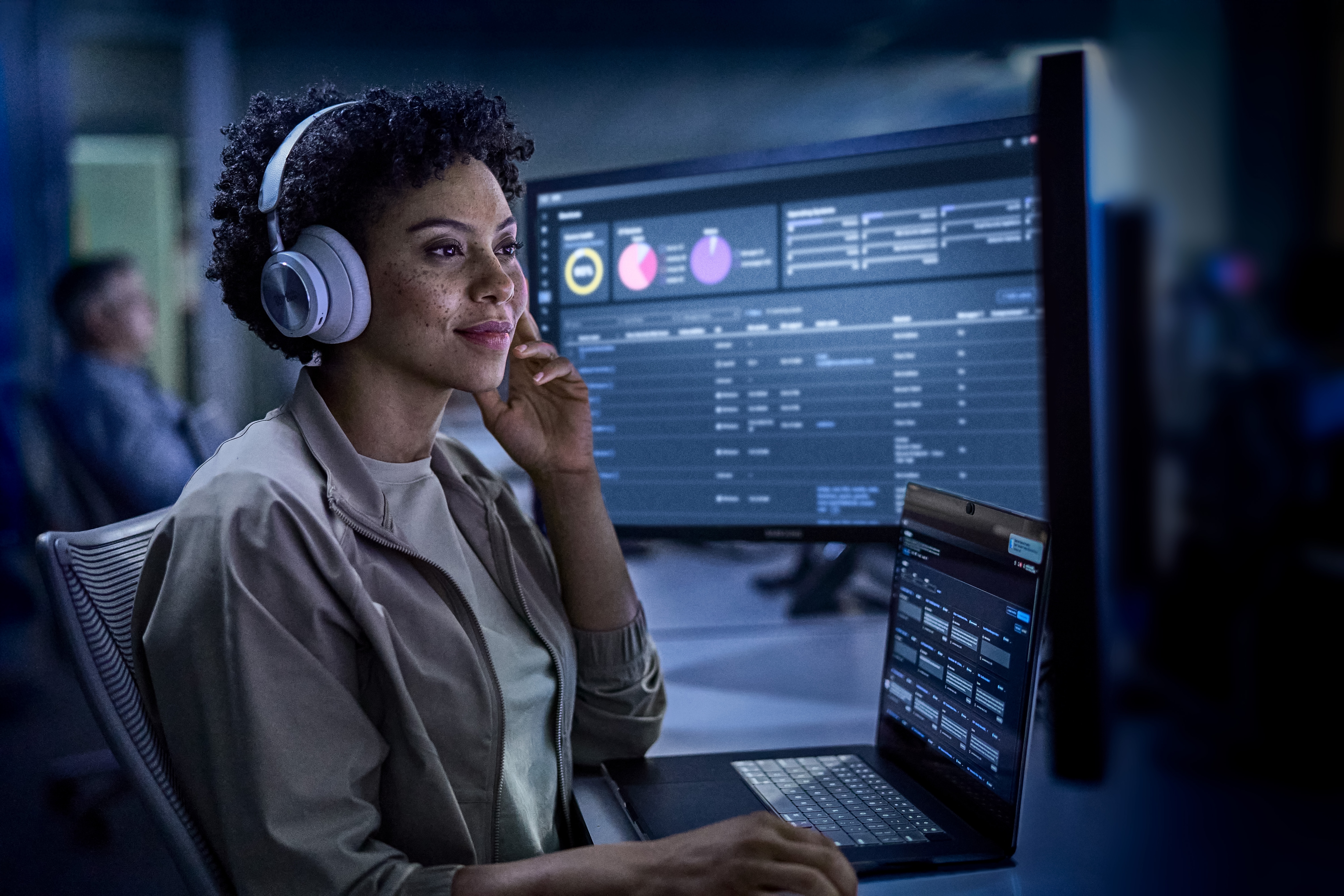 A woman wearing headphones is sitting in front of a laptop computer in a dark security operation center.