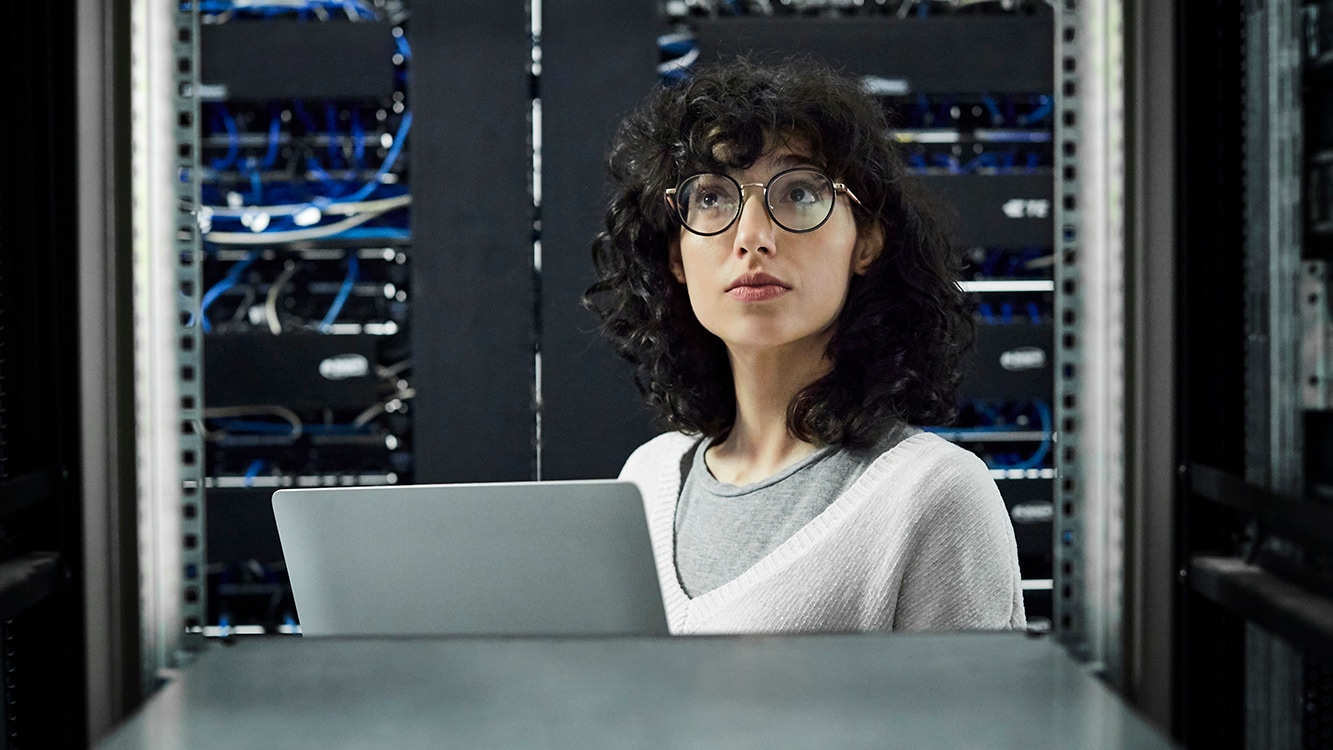 Female technician looking at cables. Engineer with laptop standing at workplace. She is in server room.